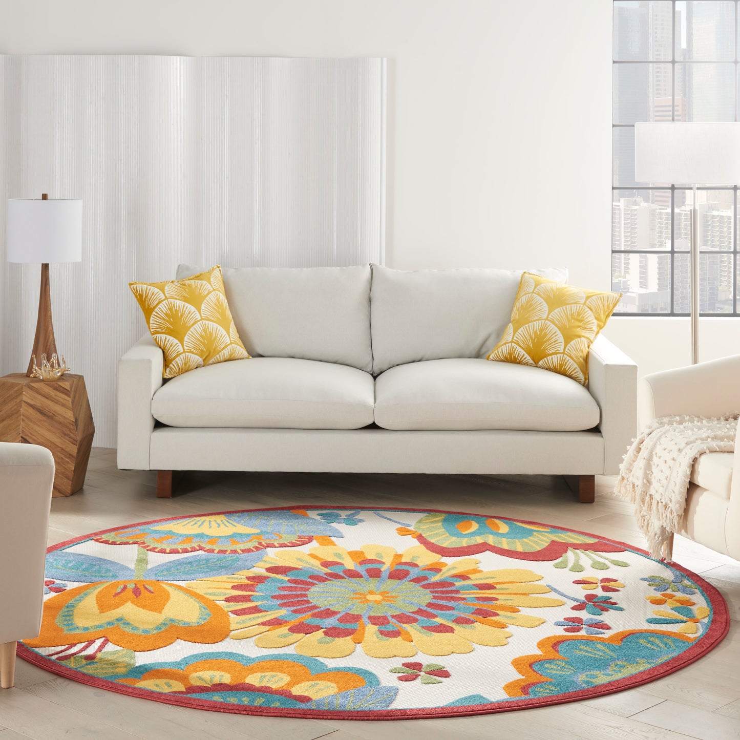 Bright living room with white sofa, yellow patterned cushions, round vibrant floral rug and city view.