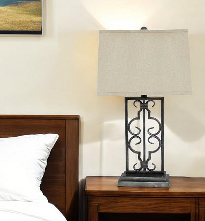 Wooden nightstand with a black wrought-iron scroll lamp and beige rectangular shade, beside a white pillow on a bed.