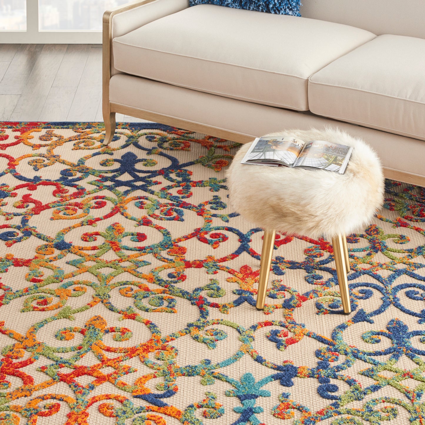 Beige sofa with light wood legs, round fluffy cream stool, and a vibrant multicolor ornate rug; magazine on stool.