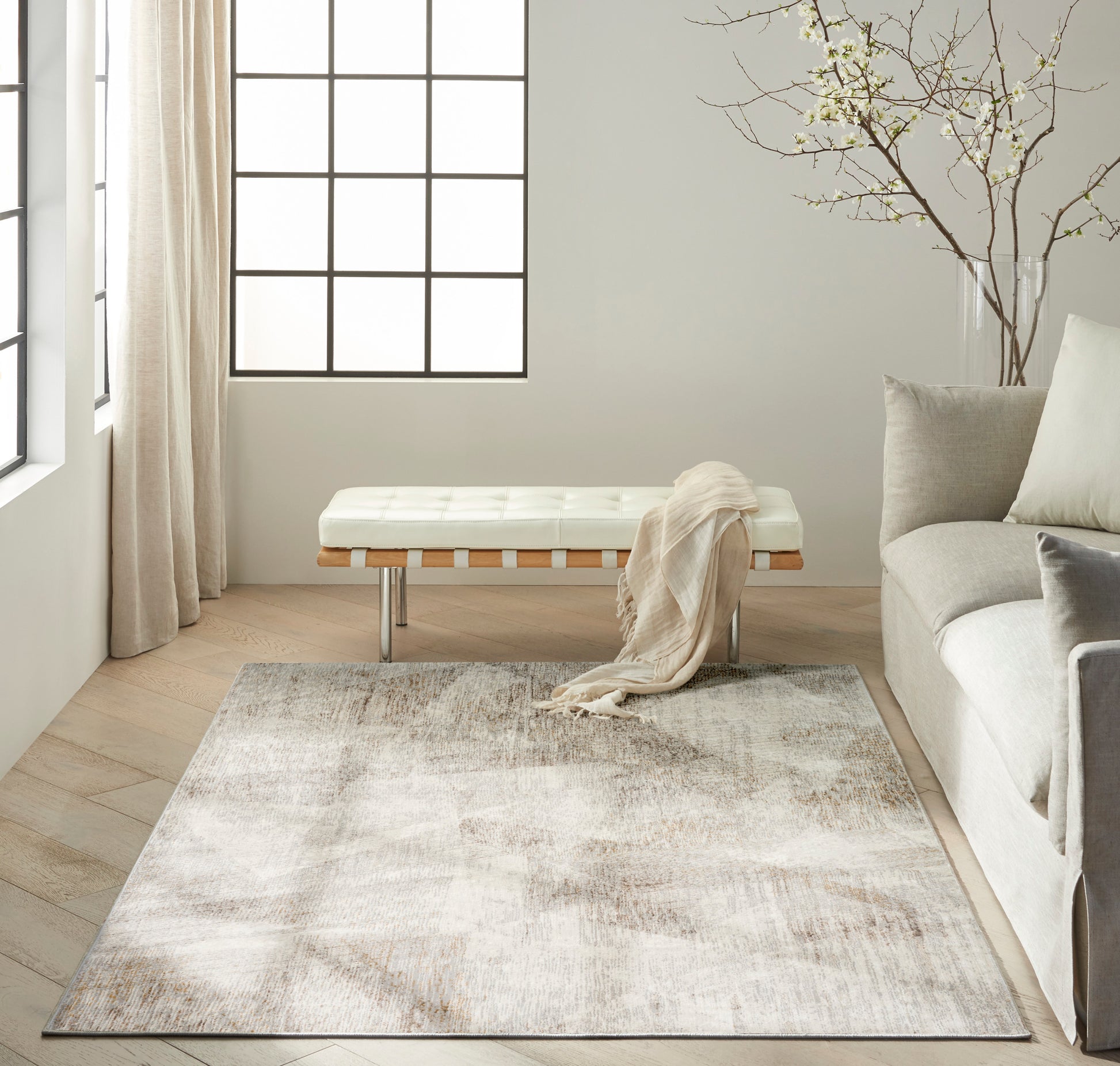 Neutral abstract area rug in beige & taupe on light wood floor, beside a white tufted bench & gray sofa in a sunlit living room.