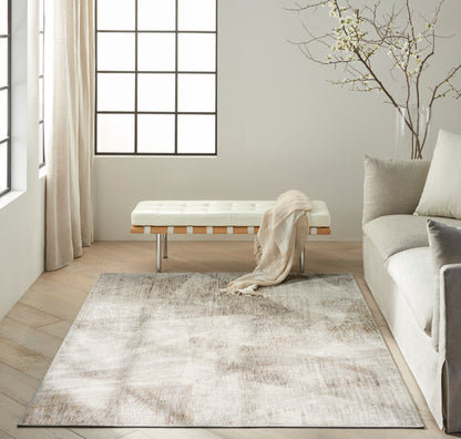 Neutral abstract area rug in beige & taupe on light wood floor, beside a white tufted bench & gray sofa in a sunlit living room.