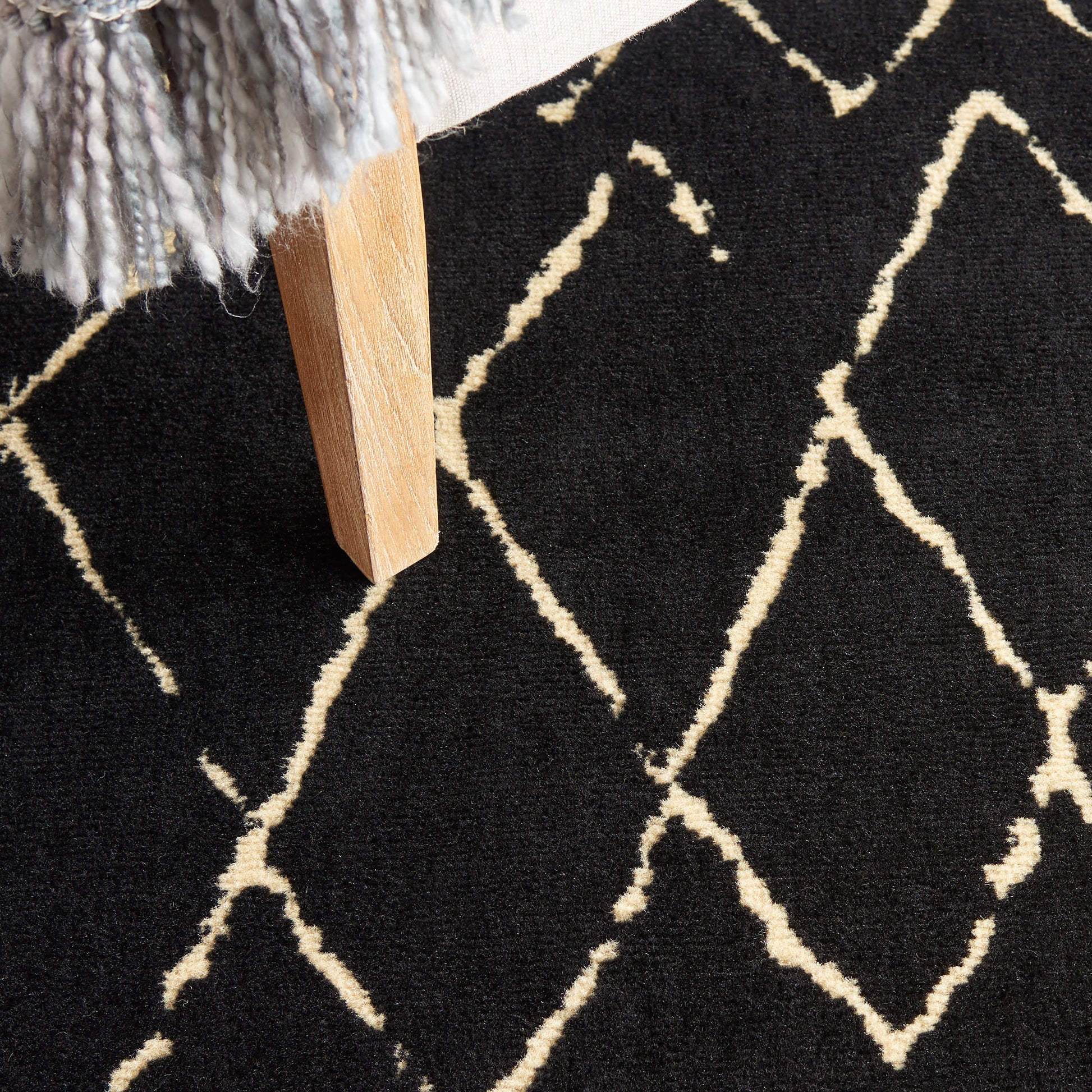 Black rug with cream geometric lattice pattern, light wood furniture leg and grey fringed throw in corner.