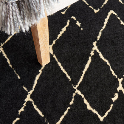 Black rug with cream geometric lattice pattern, light wood furniture leg and grey fringed throw in corner.