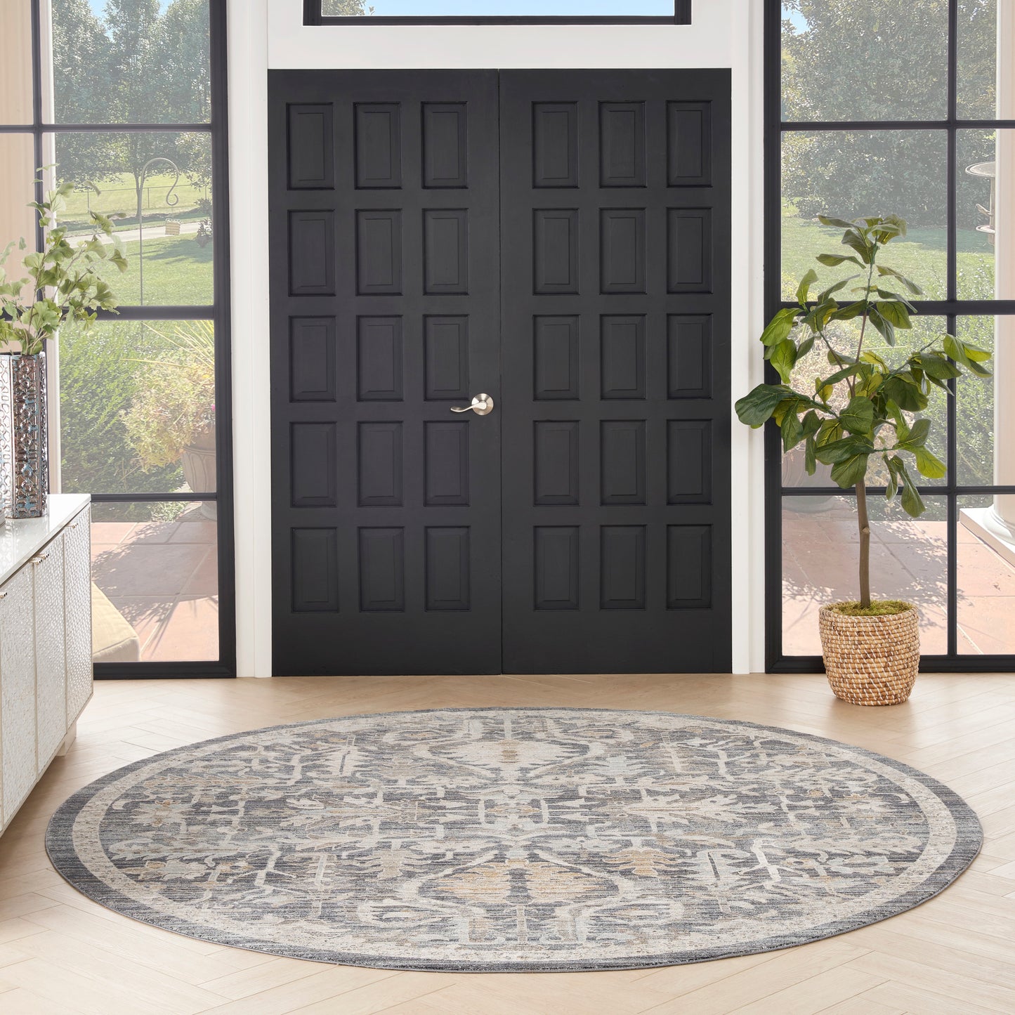Round gray patterned rug on light wood floor in a foyer with black paneled double doors and a potted plant.