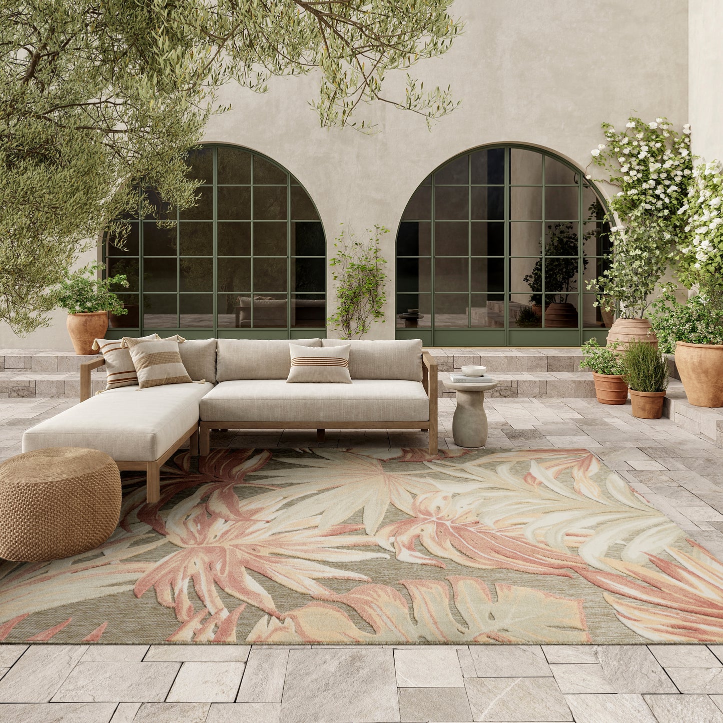 Outdoor lounge with beige sectional sofa, palm-leaf patterned rug, terracotta pots, and arched green-framed windows.