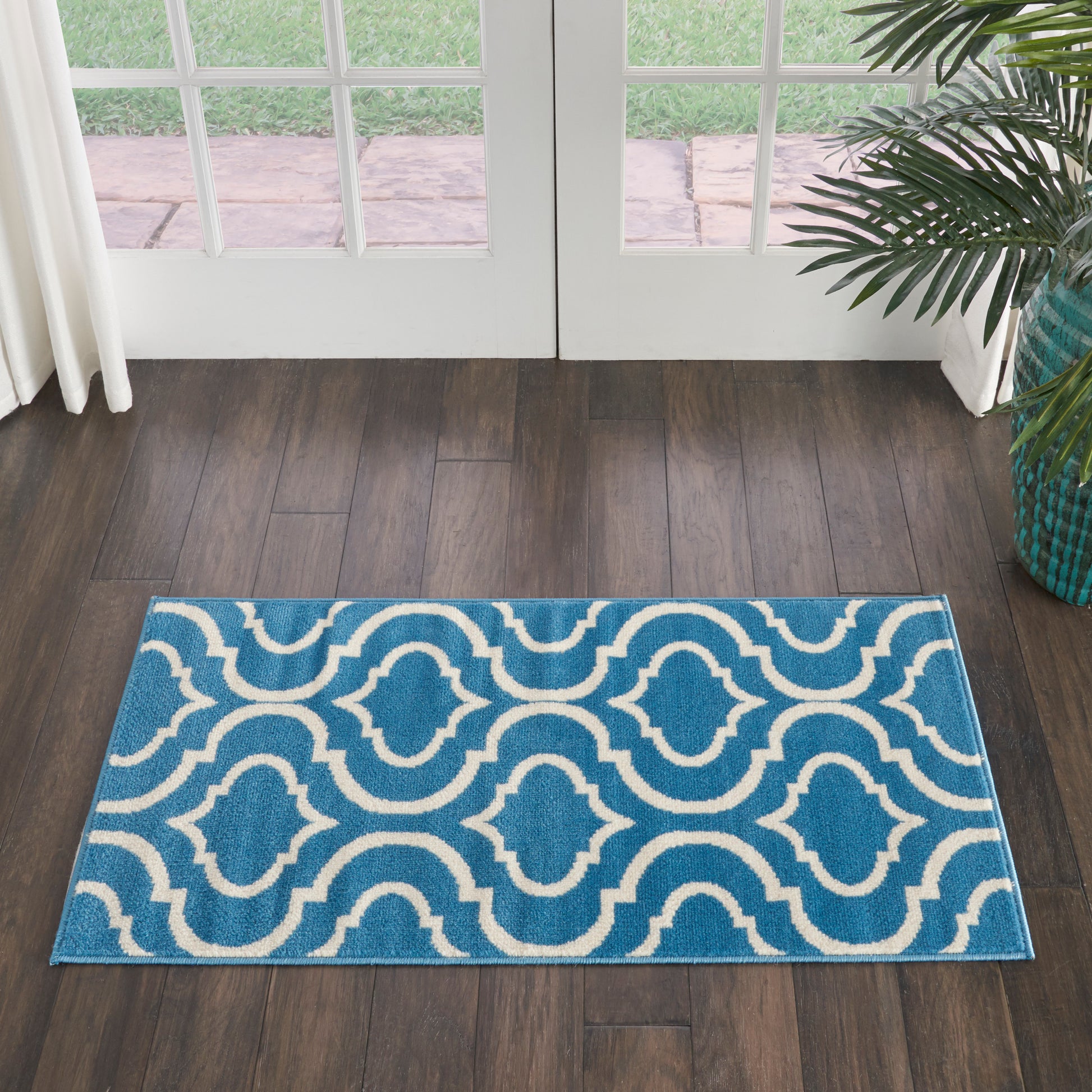 Blue rug with white interlocking wave pattern on dark wood floor in front of glass double doors; teal plant pot nearby.