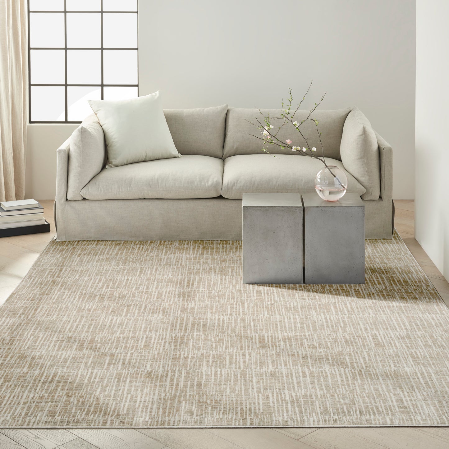 Beige fabric sofa with pale cushions, gray concrete cube side tables, glass vase with blossoms, beige textured rug.