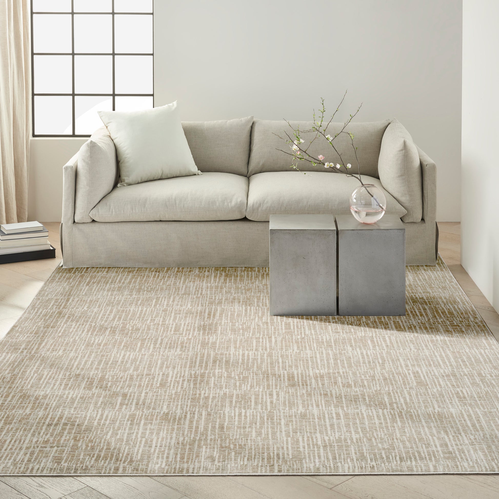 Beige fabric sofa with pale cushions, gray concrete cube side tables, glass vase with blossoms, beige textured rug.
