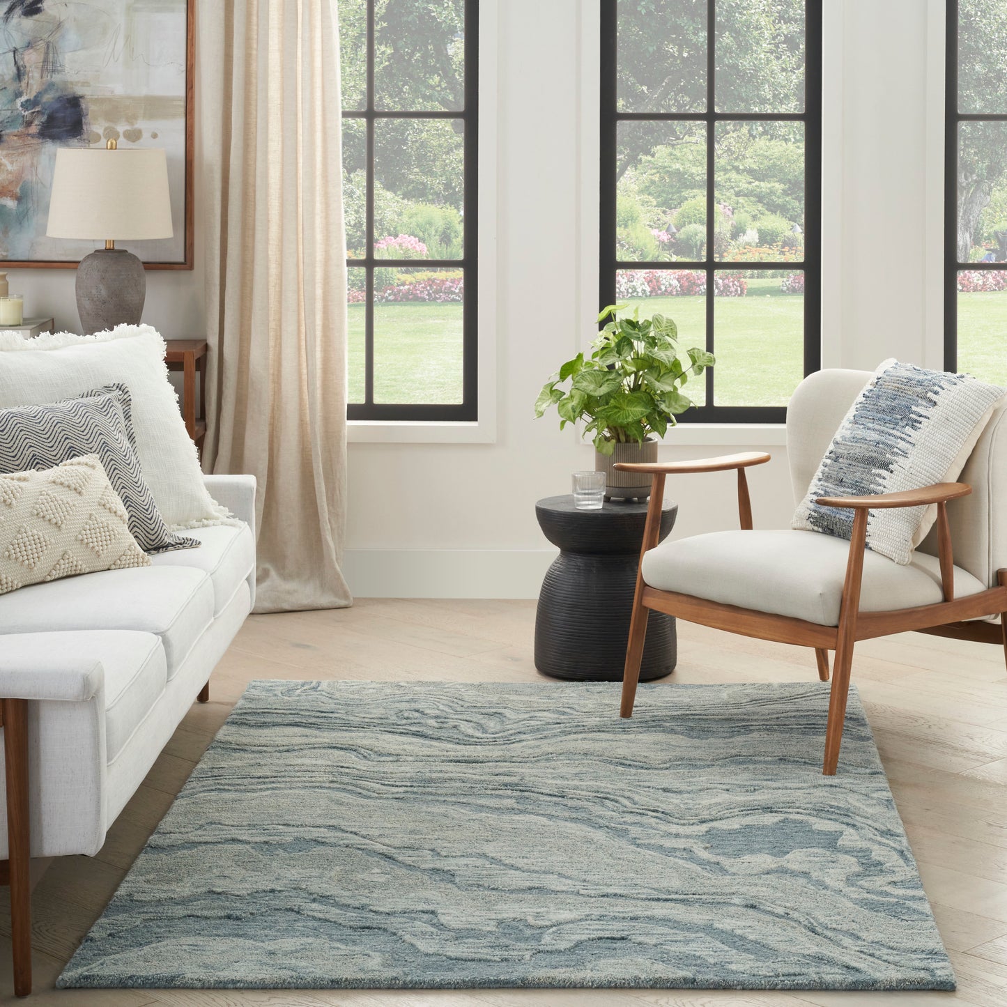 Bright living room with cream sofa, wooden-armchair, blue wavy-area rug, potted plant, black side table near grid windows.
