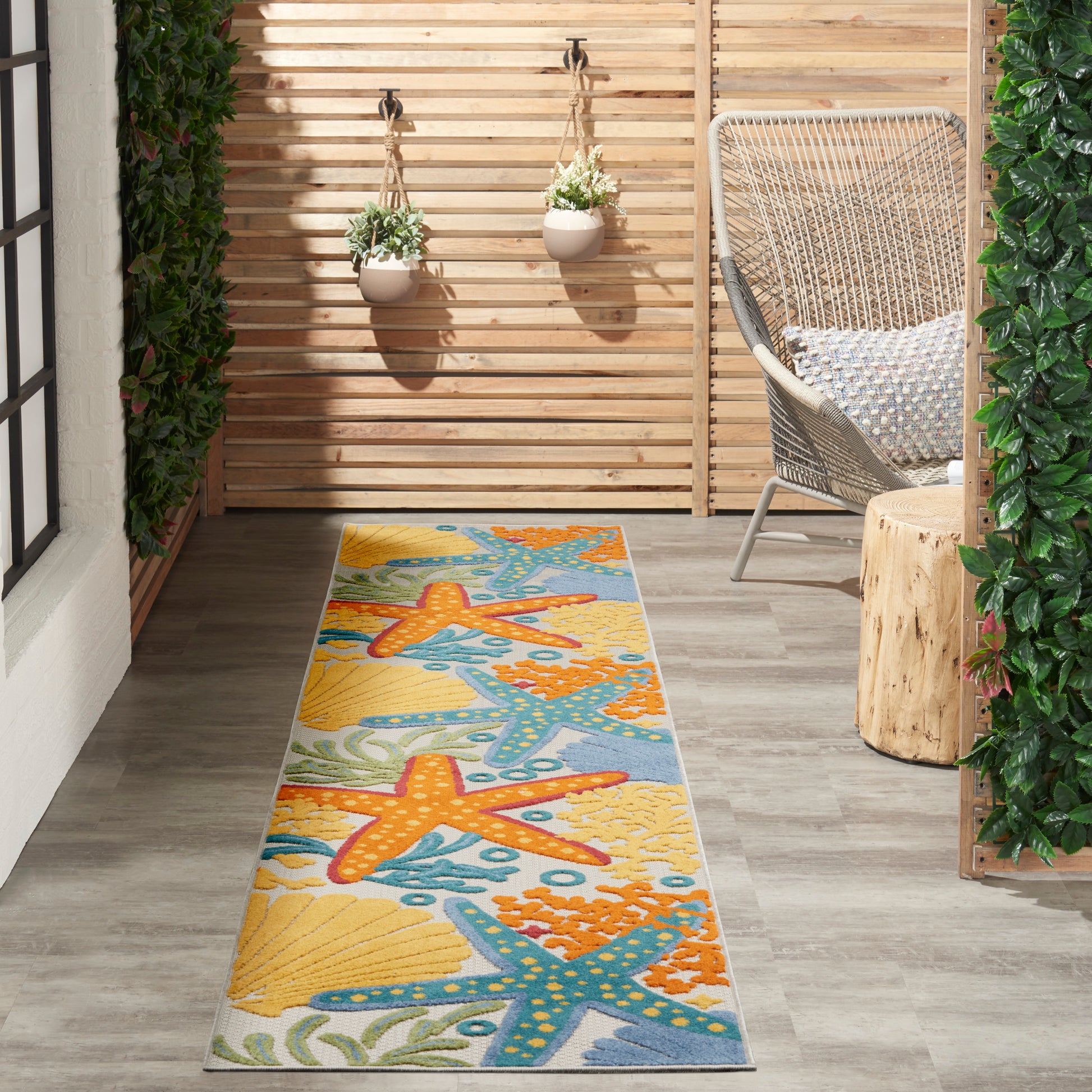 Outdoor runner rug with orange starfish & sea life on yellow/blue background, laid on a wooden deck beside a wicker chair.