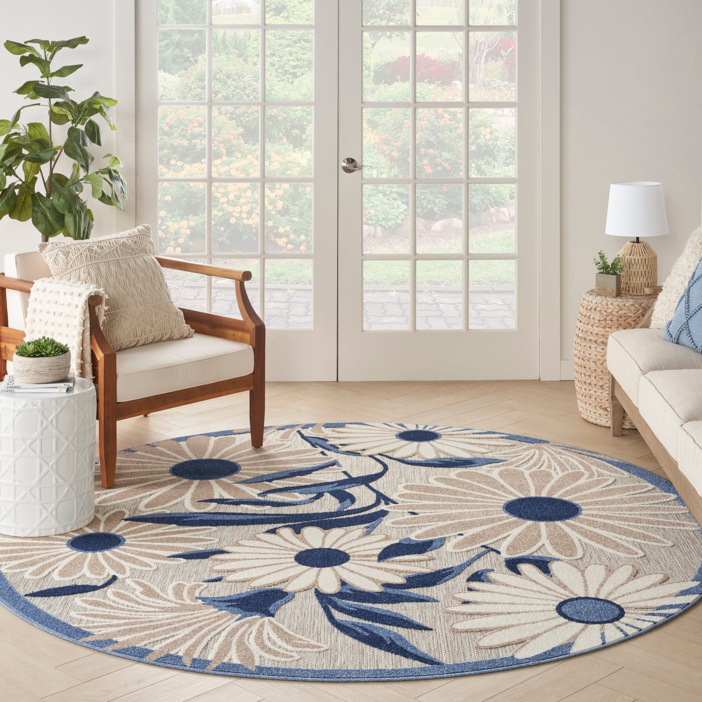 Round beige rug with large cream daisy motifs and navy blue accents in a sunlit living room by French doors.