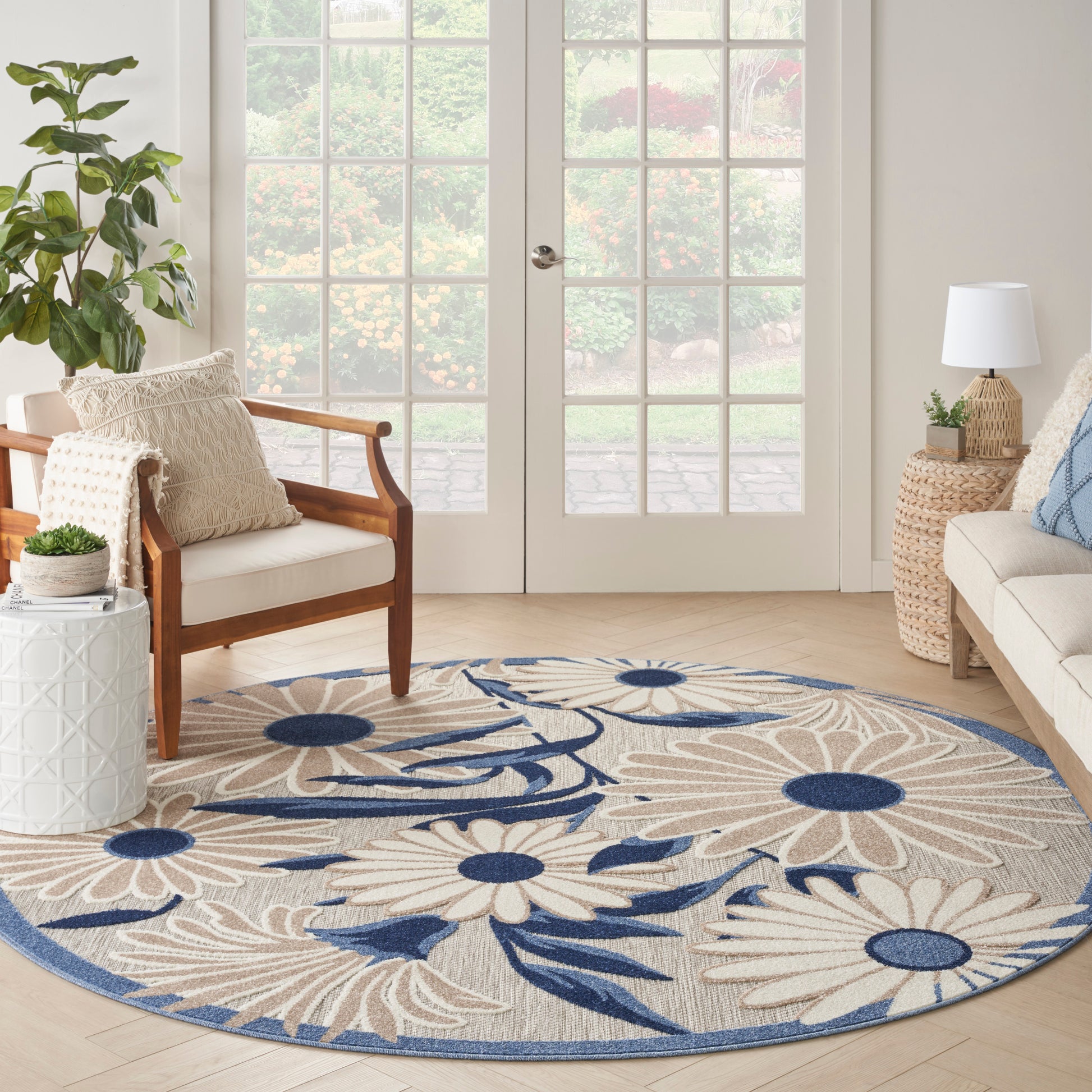 Round beige rug with large cream daisy motifs and navy blue accents in a sunlit living room by French doors.