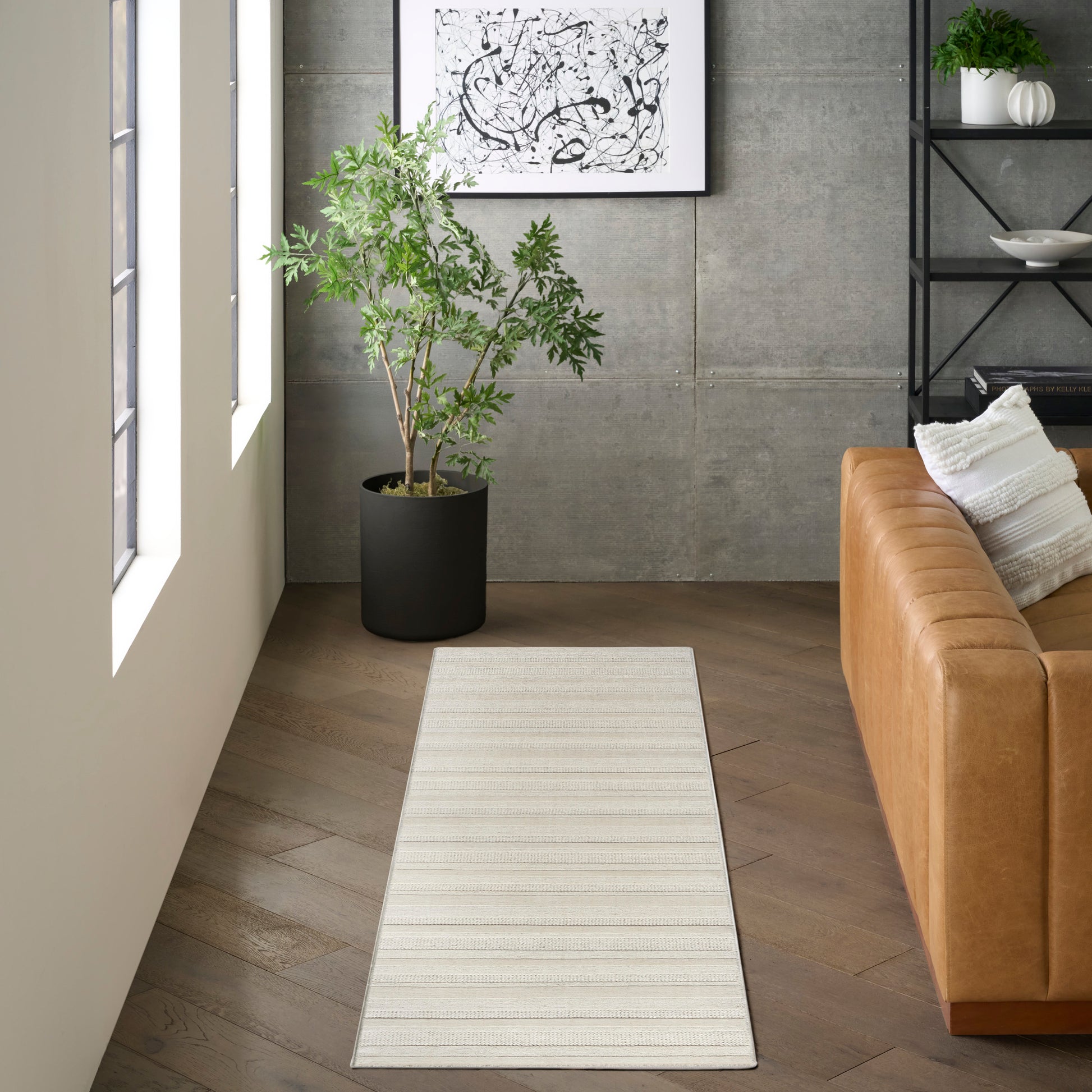 Beige striped runner rug on wood floor beside a tan leather sofa; potted plant and abstract art on a concrete wall.