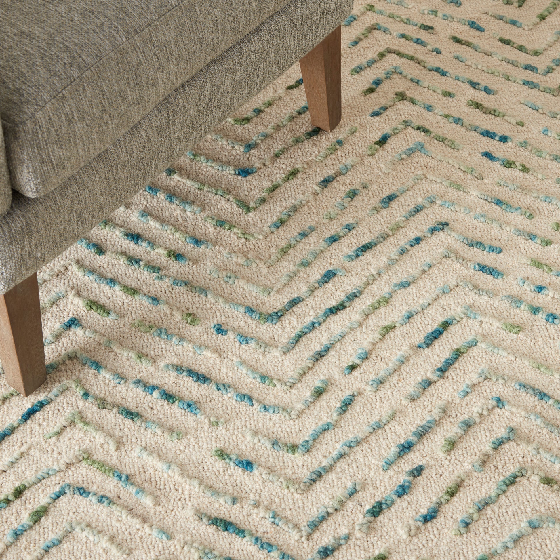 Beige looped-pile rug with blue, teal & green chevron accents under a gray sofa with wooden legs.