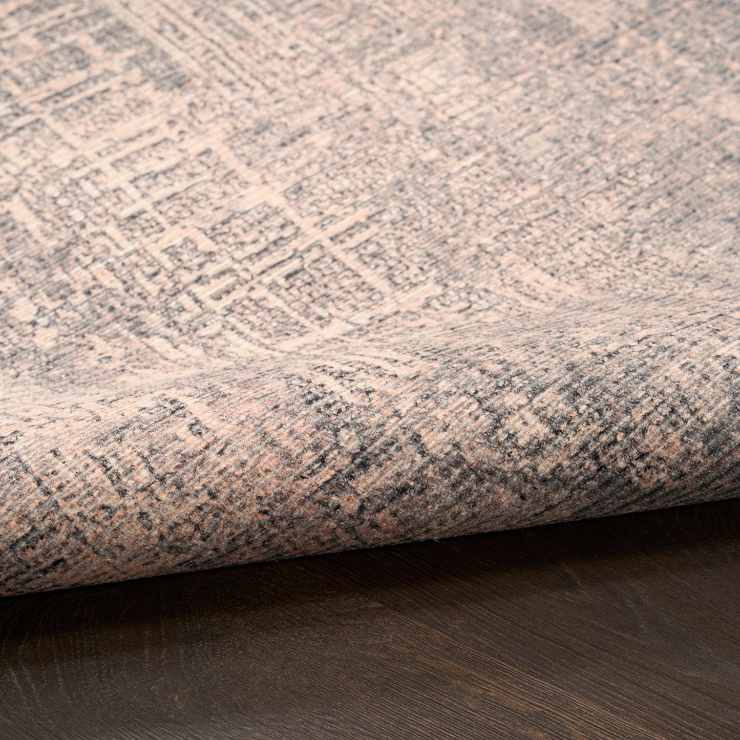 Close-up of a beige-gray woven rug with a distressed geometric pattern, folded at the edge on a dark wood floor.