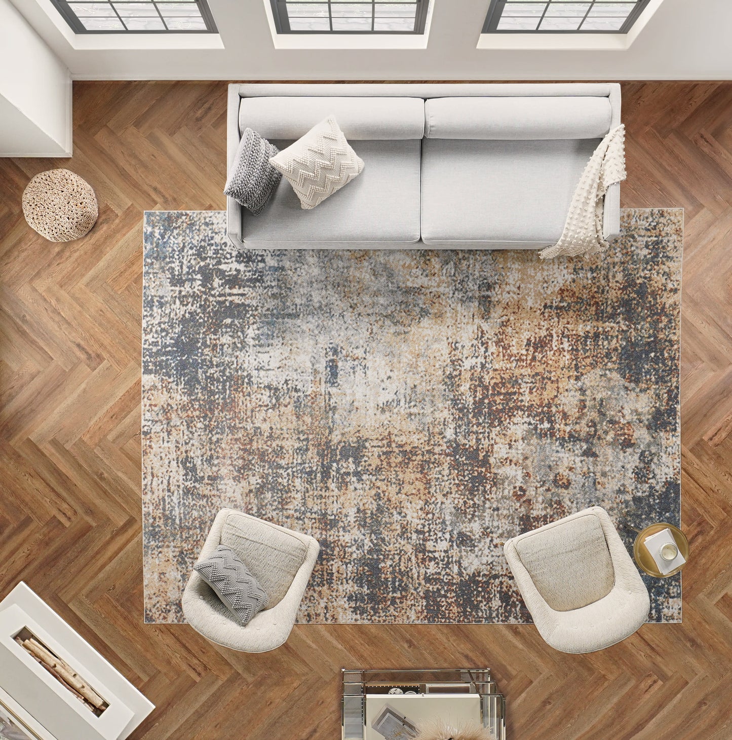 Top-down view of modern living room: blue-gray abstract rug on herringbone wood floor, light gray sofa, cream chairs, pouf.