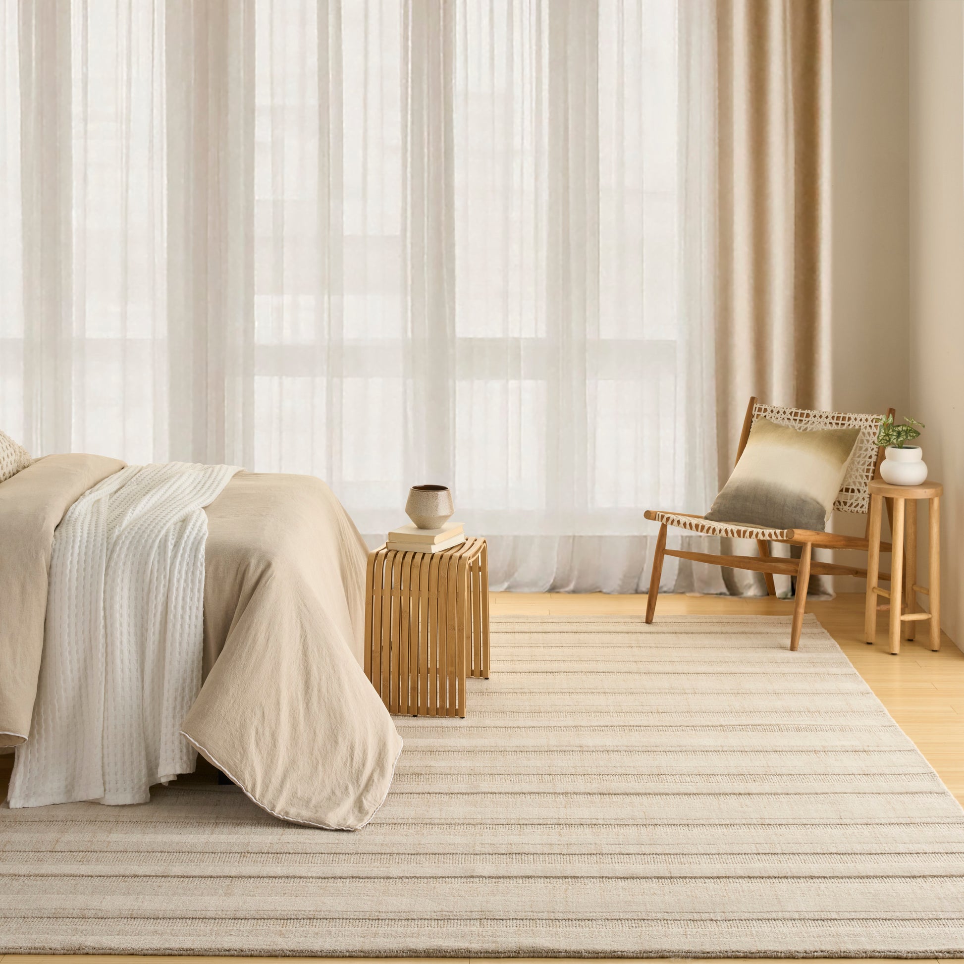Neutral beige bedroom with layered bedding, striped rug, woven chair, small plant on a wood table, by sheer curtains.