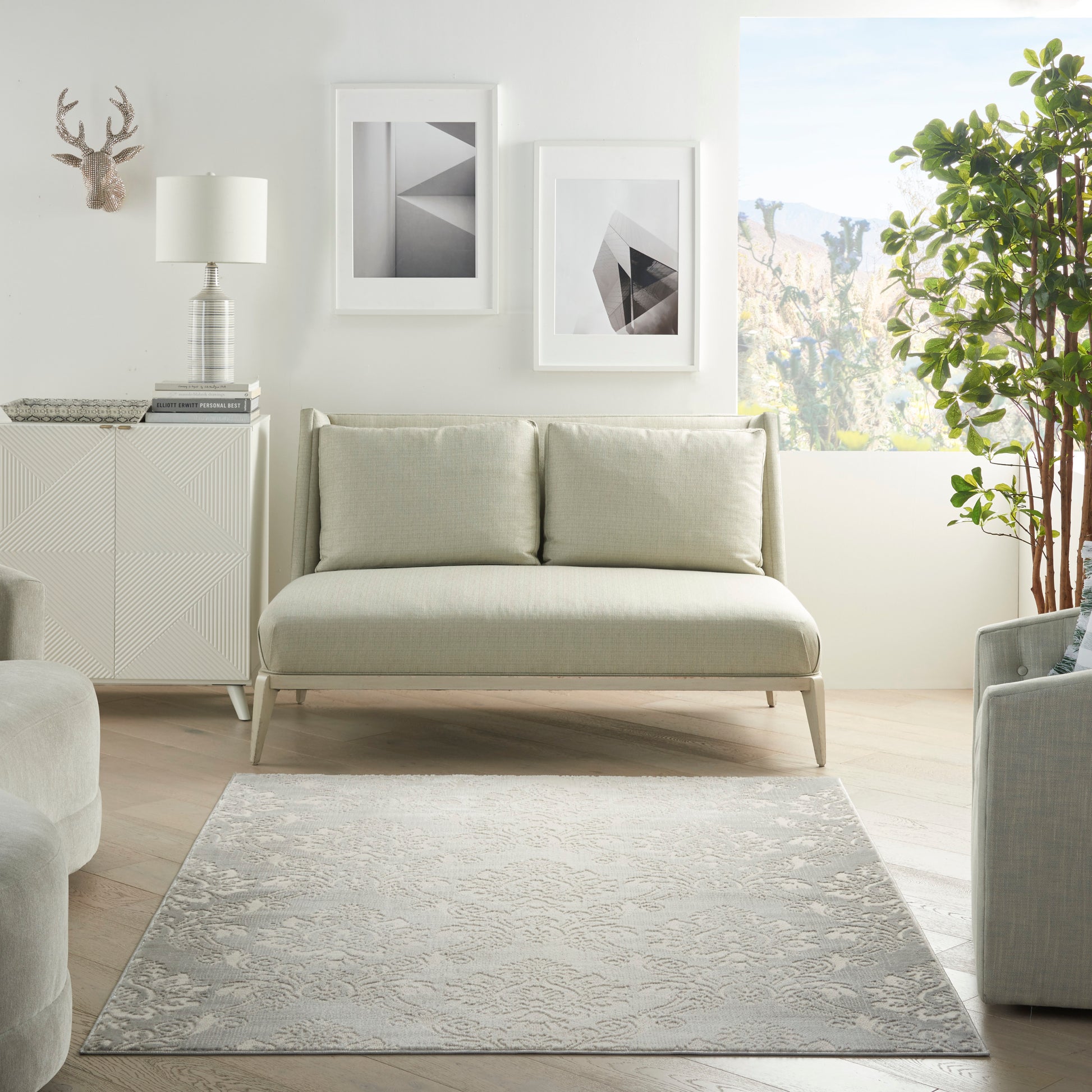 Modern neutral living room: beige fabric sofa, light wood legs, gray patterned rug, white framed art, leafy plant by window.