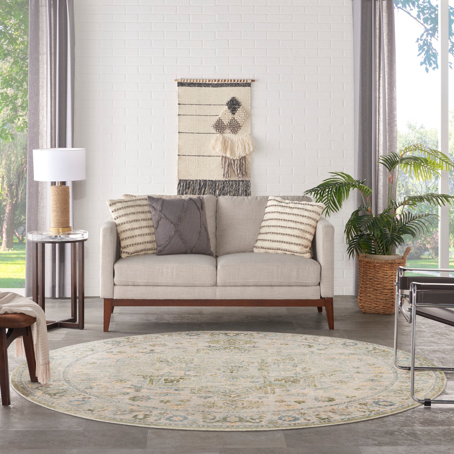 Beige fabric sofa with wooden legs, gray and striped pillows, round patterned rug, woven wall hanging, potted plant.