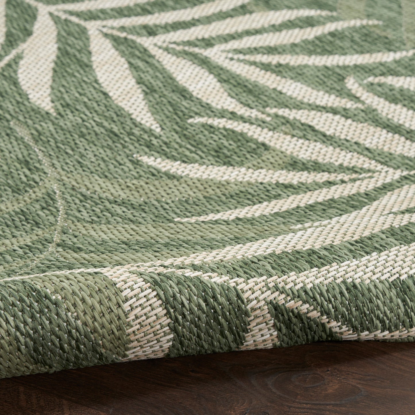 Green & cream outdoor rug with a leafy botanical pattern, flat-weave texture, rolled edge on a dark wood floor, size 5' x 7'.