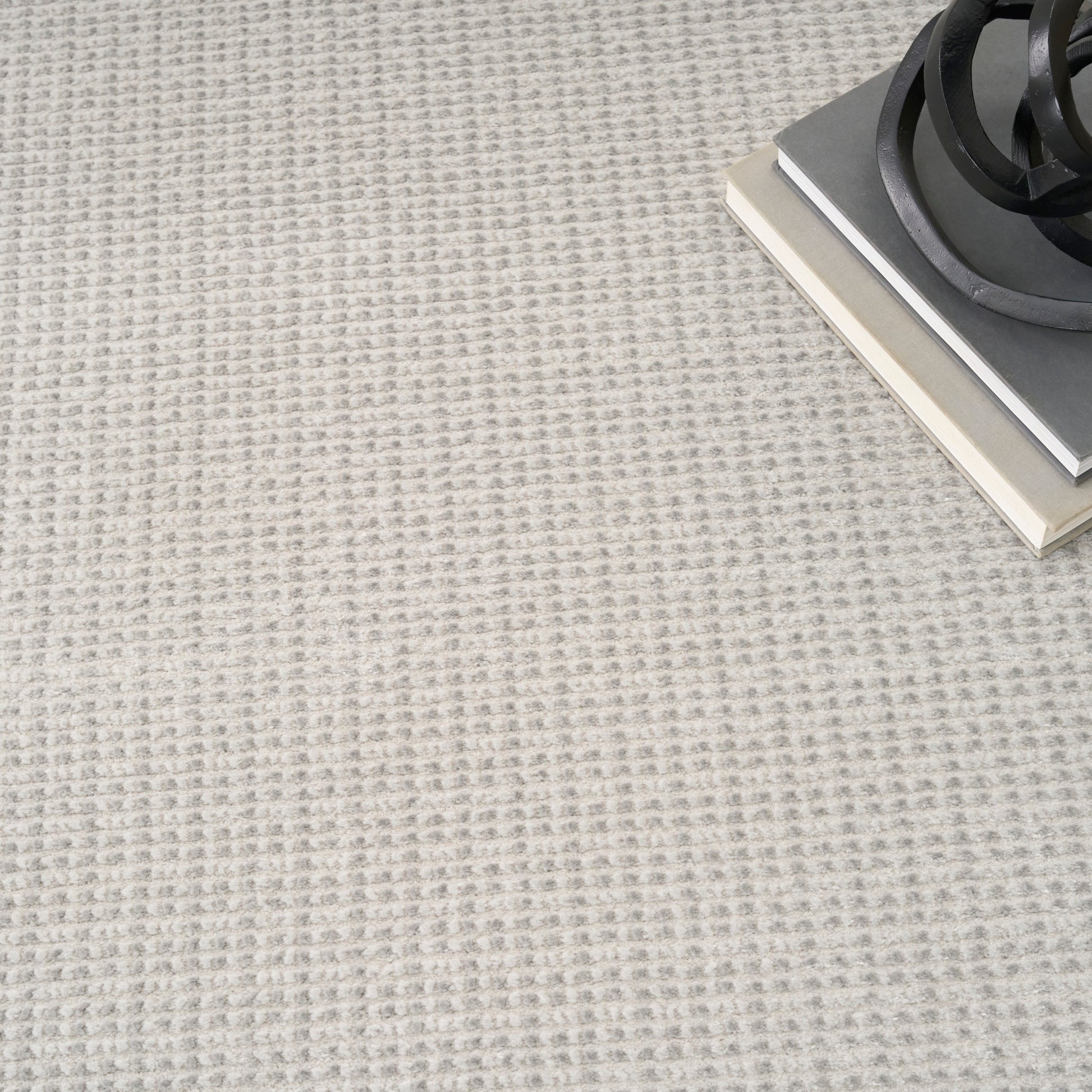 Beige textured rug with subtle square pattern; top-right shows a stack of gray books and a black spiral metal sculpture.