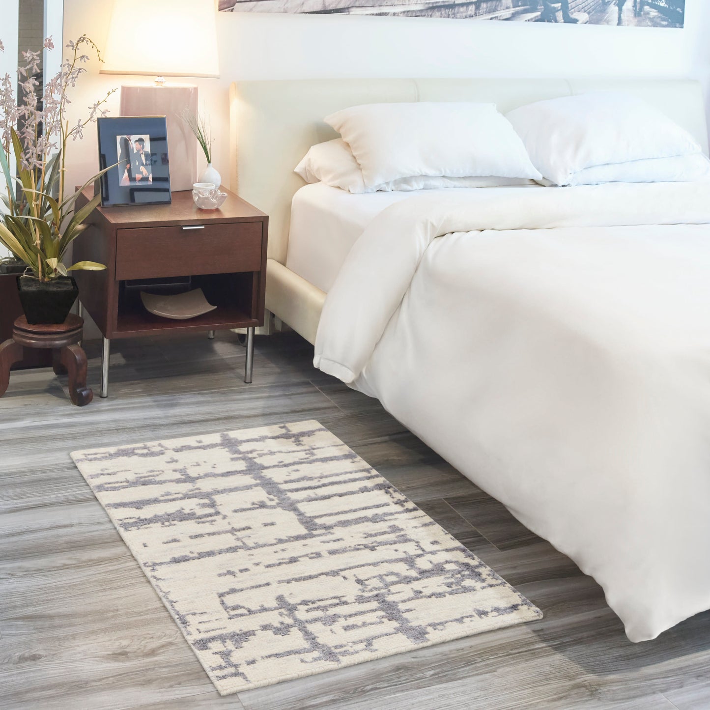 White bedding with a wooden nightstand, lamp, framed photo, potted plant; gray geometric rug on wood floor.