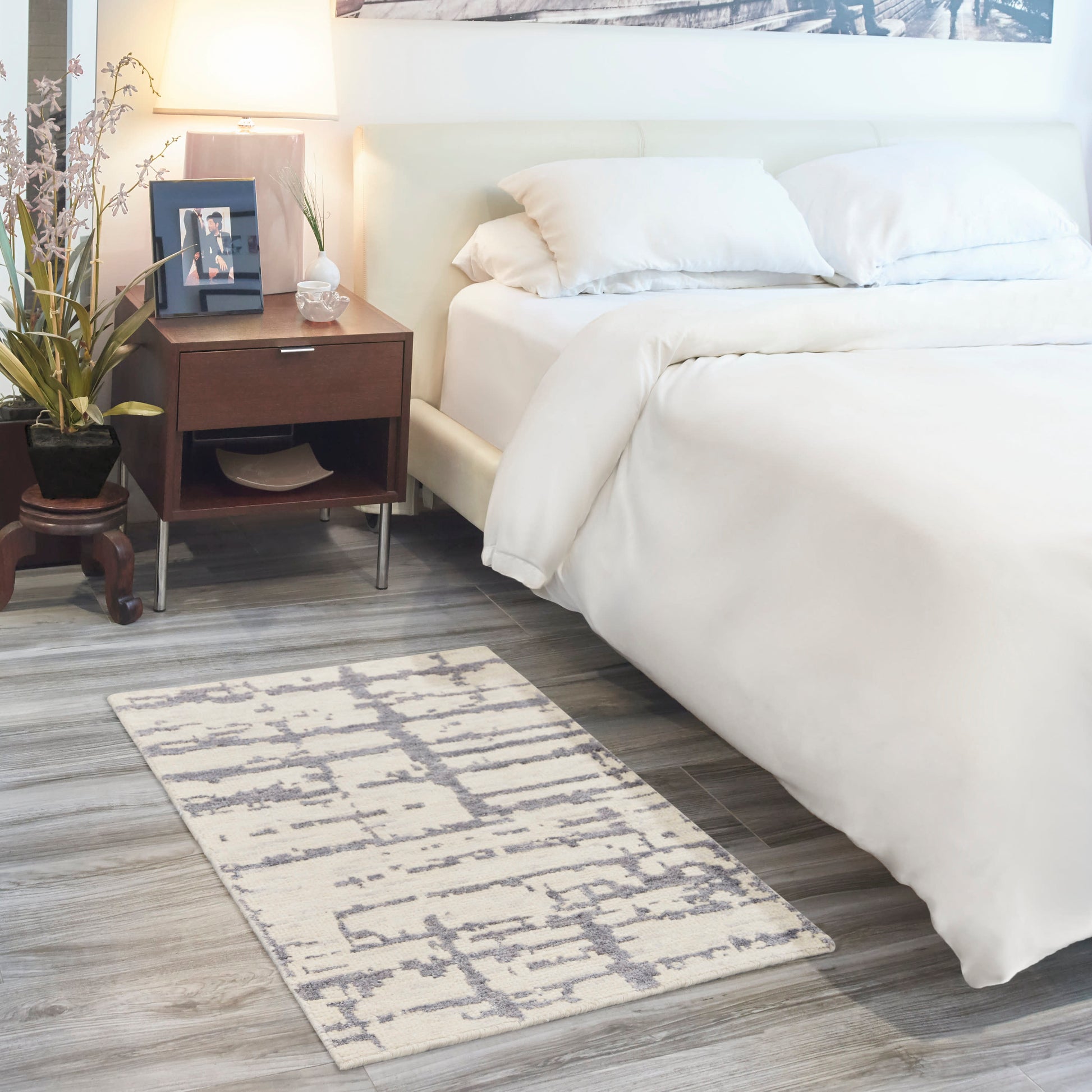 White bedding with a wooden nightstand, lamp, framed photo, potted plant; gray geometric rug on wood floor.
