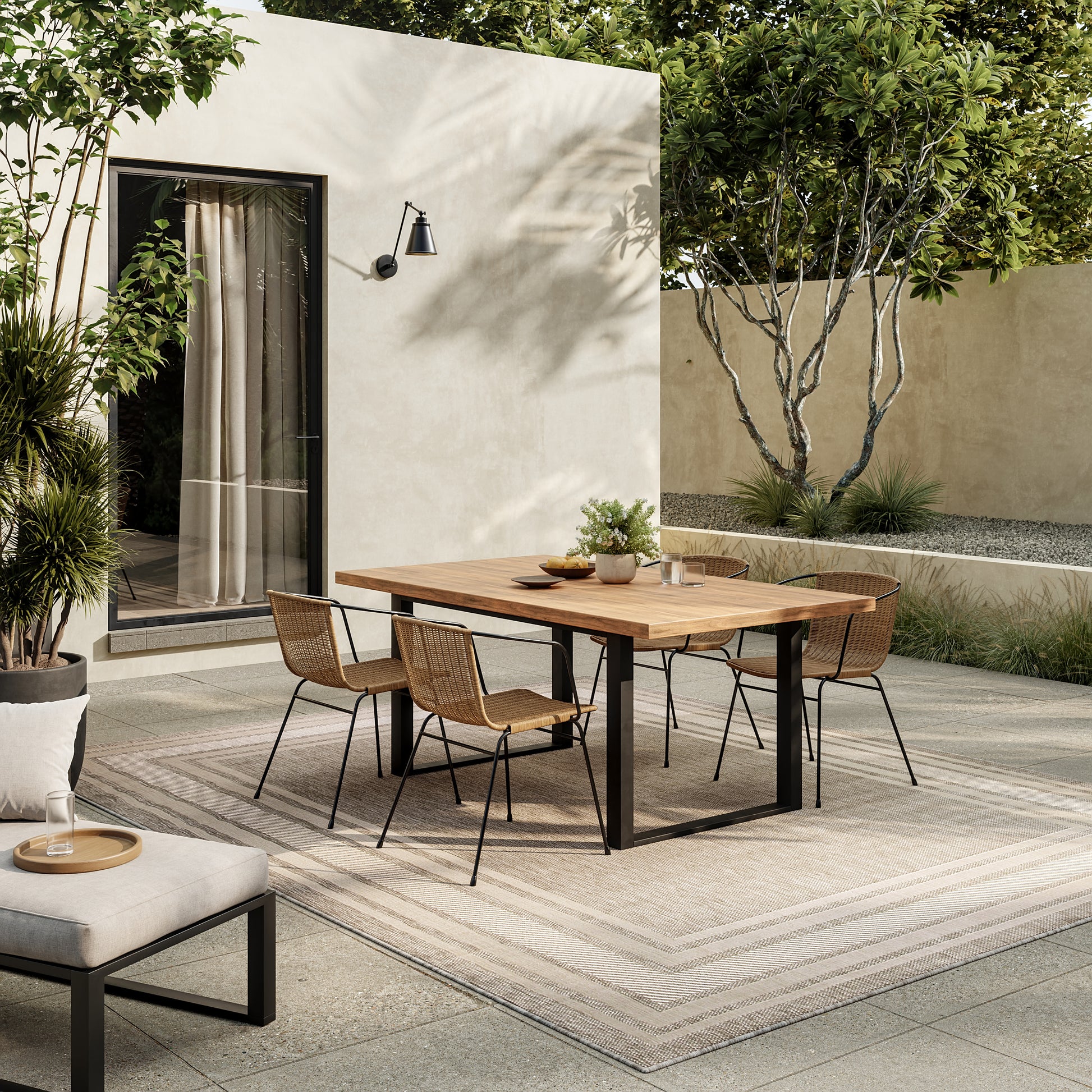 Outdoor dining area with rectangular wooden table and black metal legs, six wicker chairs, on a light rug by beige walls and greenery.