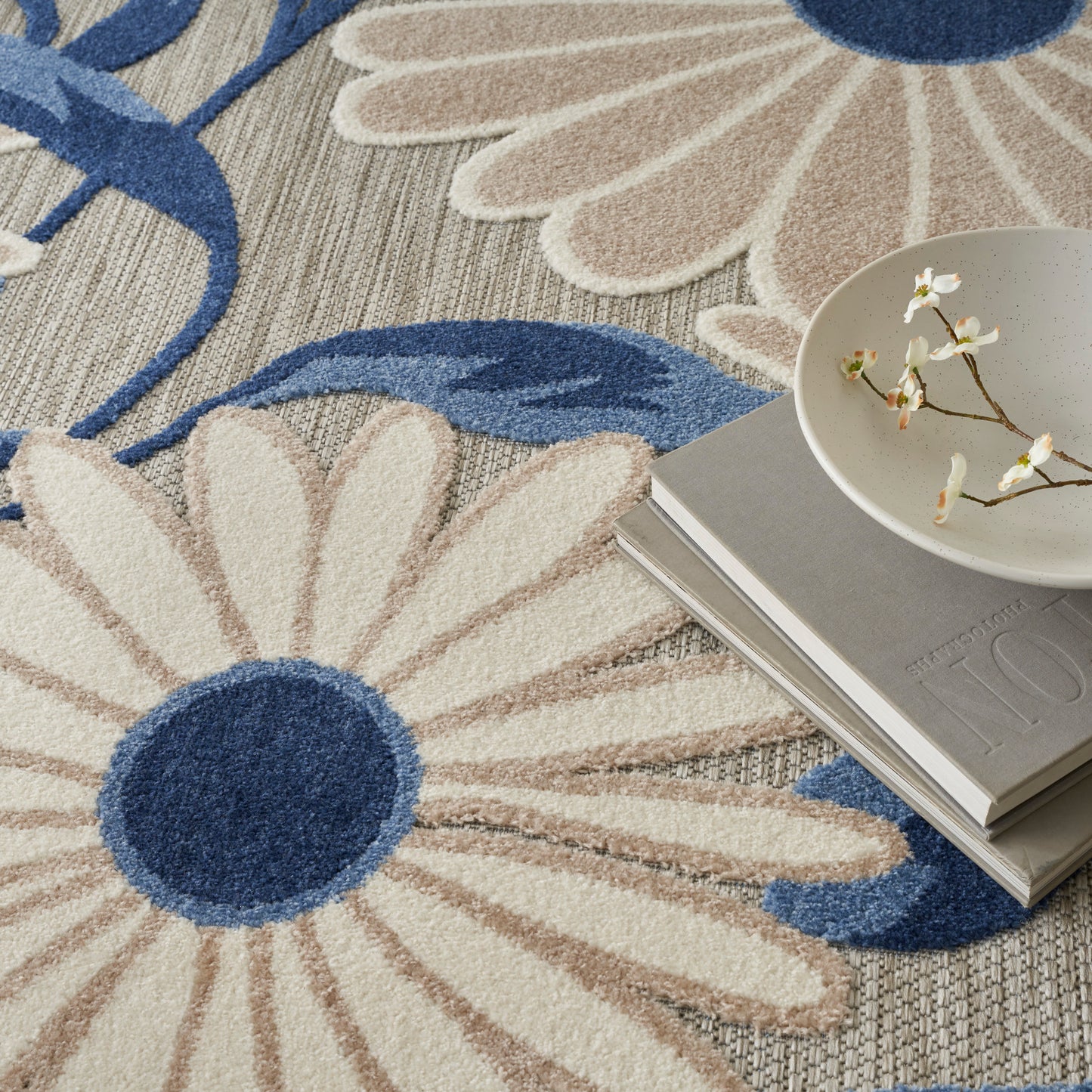 Beige and blue floral rug with large daisy motifs, beside stacked gray books and a white ceramic bowl with blossoms.