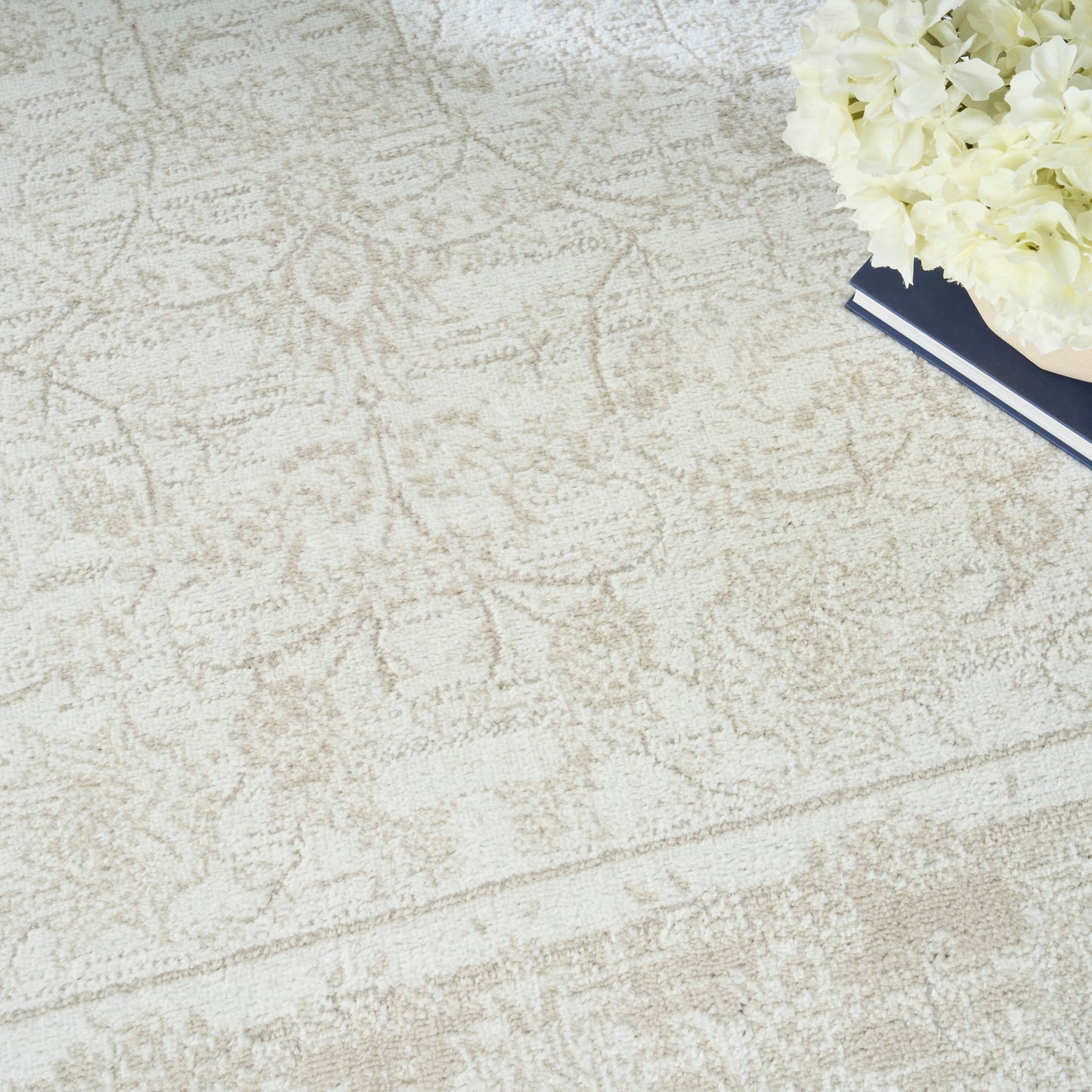 Beige patterned area rug with a damask motif; white hydrangea bouquet atop navy-blue books in the corner.