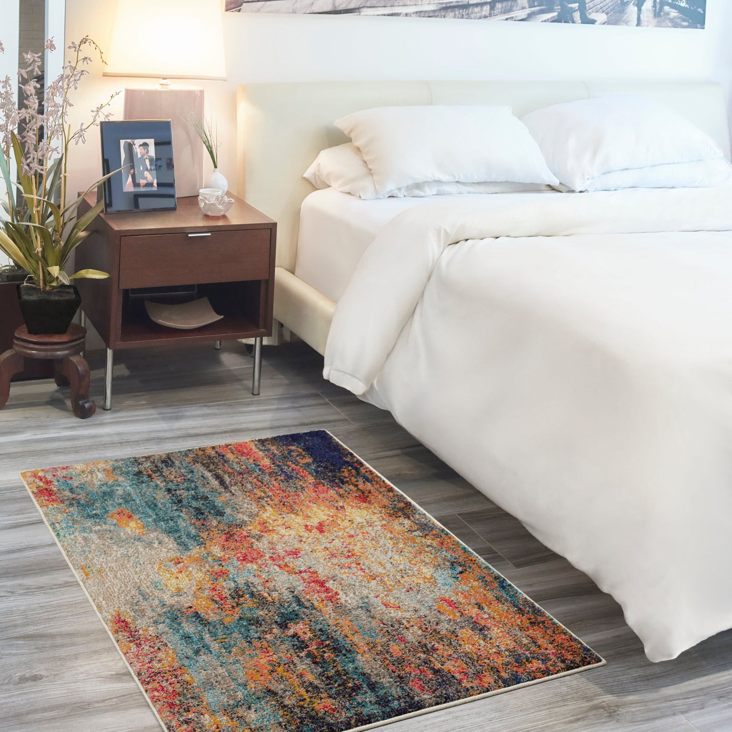 Abstract multicolor area rug on gray wood floor beside white bed with dark wood nightstand and lamp.