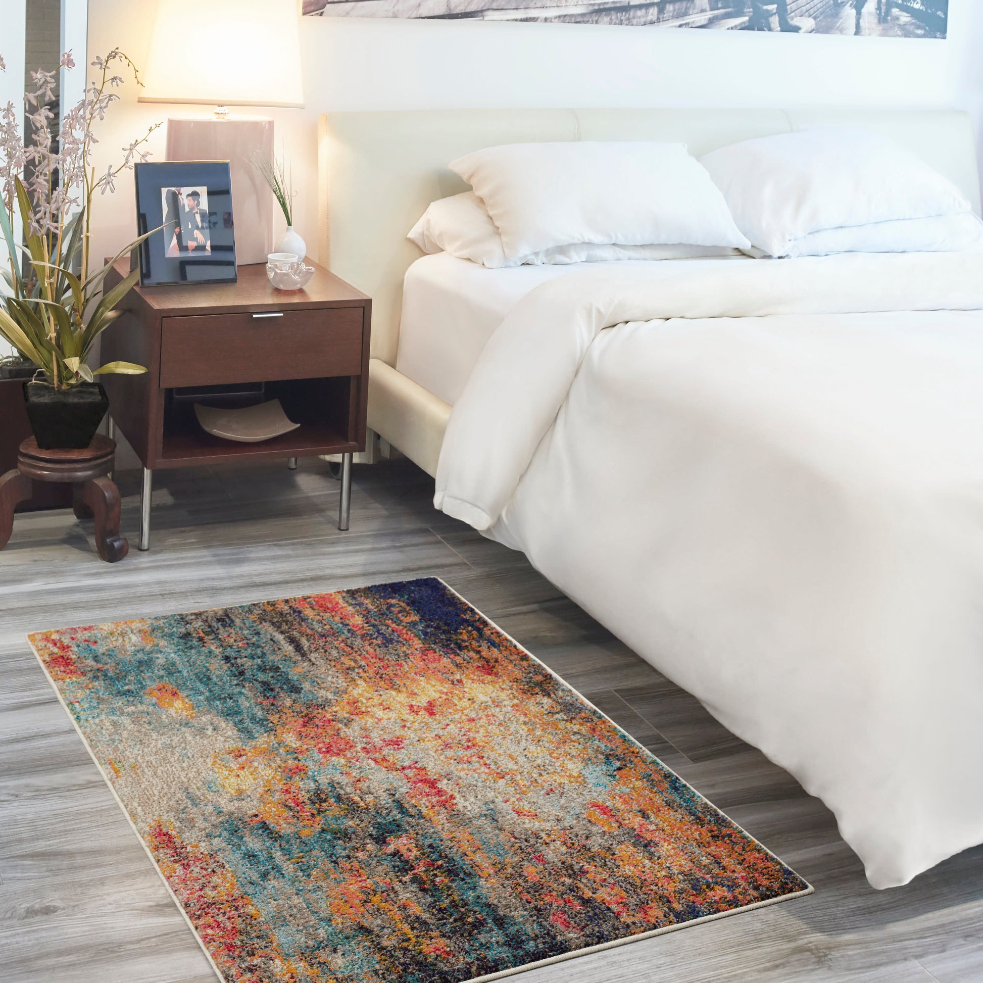 Abstract multicolor area rug on gray wood floor beside white bed with dark wood nightstand and lamp.