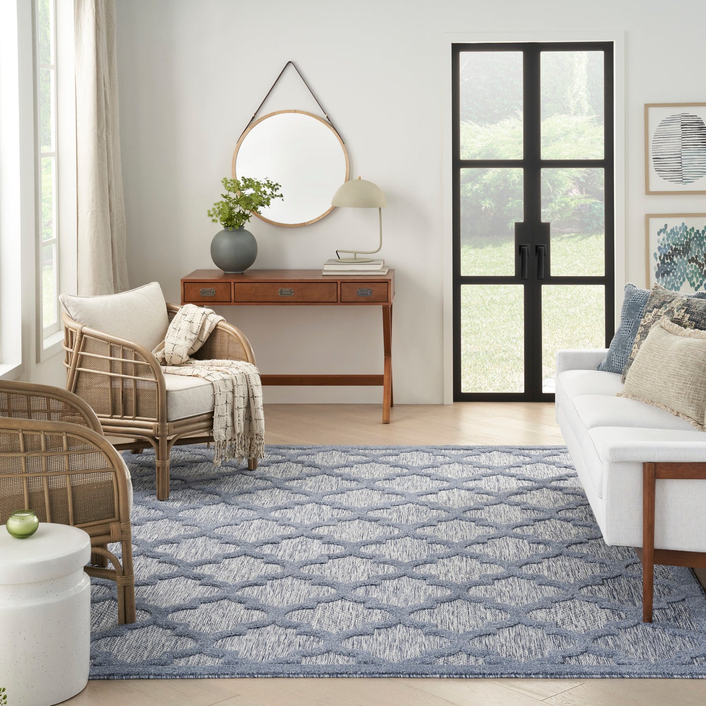 Bright living room with rattan chairs, white sofa, blue geometric rug, console with round mirror, plant, black-framed glass door.