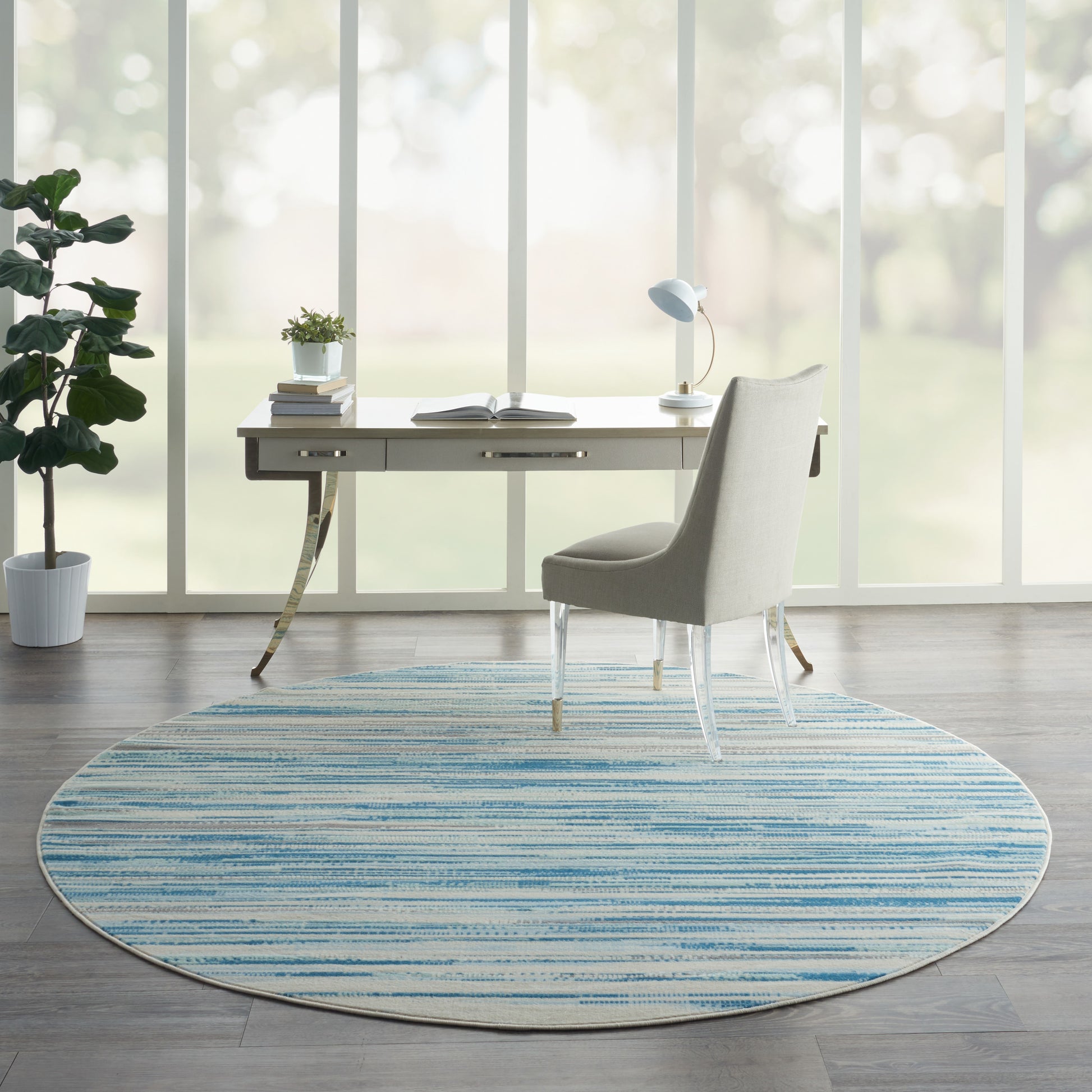 Modern home office with circular blue striped rug, light-wood desk, light gray upholstered chair, lamp and plants by windows.