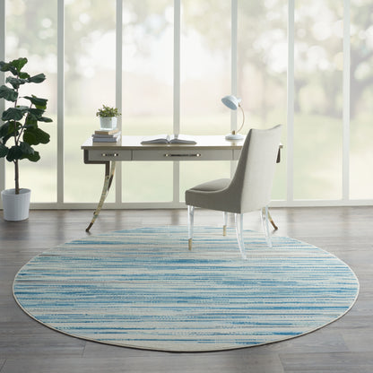 Modern home office with circular blue striped rug, light-wood desk, light gray upholstered chair, lamp and plants by windows.