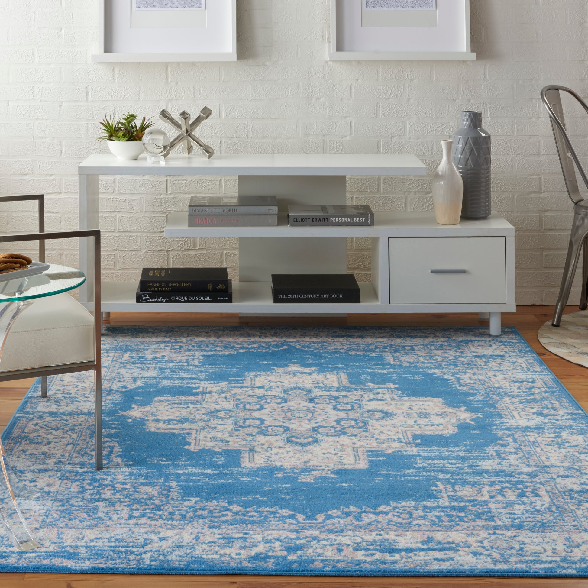 White modern console with books, vases, and sculpture on a white brick wall, blue distressed rug beneath.