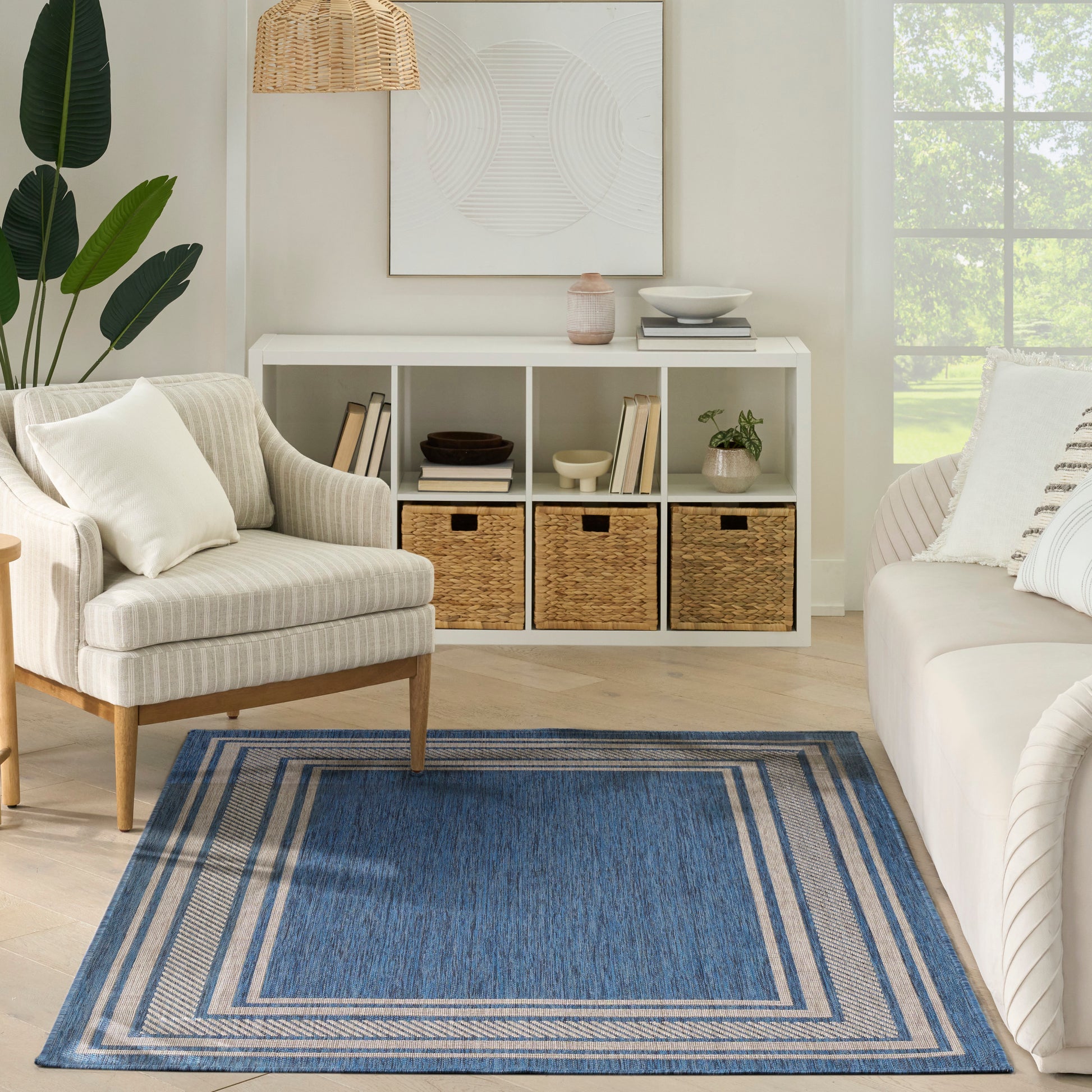 Neutral living room with white cubby storage, wicker baskets, beige armchair, light sofa, blue bordered rug, plant.