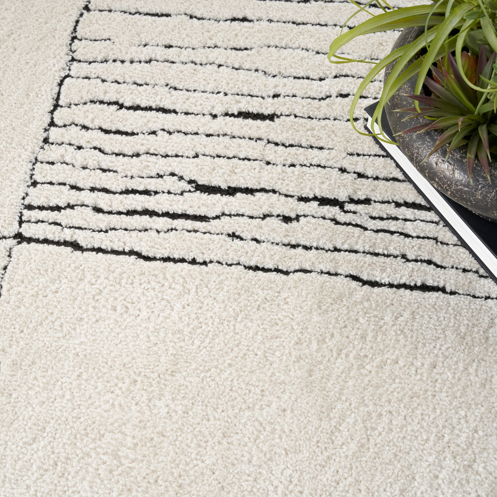 Cream shag rug with black abstract stripes; plush pile, modern minimalist decor beside a potted plant.