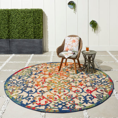 Round multicolor ornate rug on a patio with wicker chair, side table, cushion, glass of drink, white wall and greenery.