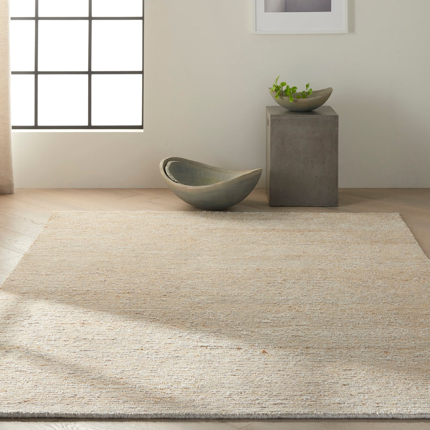 Natural jute rug in a minimalist room with a curved gray-green ceramic bowl and a concrete pedestal with a small plant.