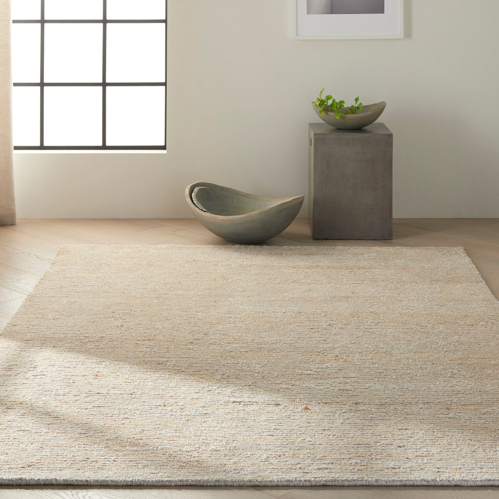 Natural jute rug in a minimalist room with a curved gray-green ceramic bowl and a concrete pedestal with a small plant.