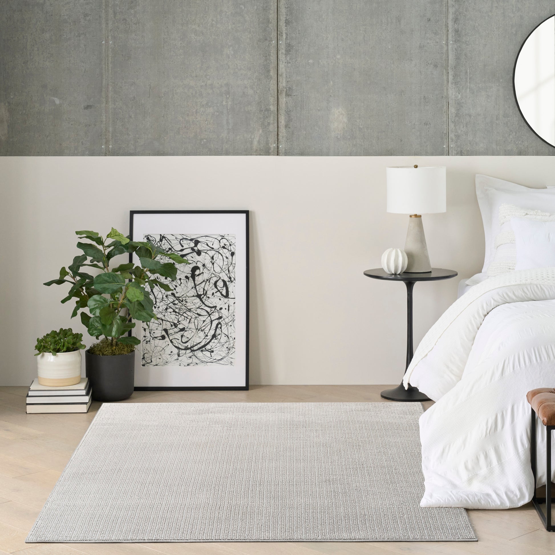 Modern bedroom corner with white bedding, black-framed abstract art, potted plant, books, gray area rug, and round mirror.