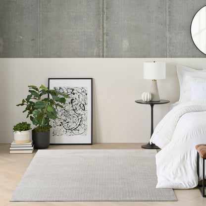 Modern bedroom corner with white bedding, black-framed abstract art, potted plant, books, gray area rug, and round mirror.