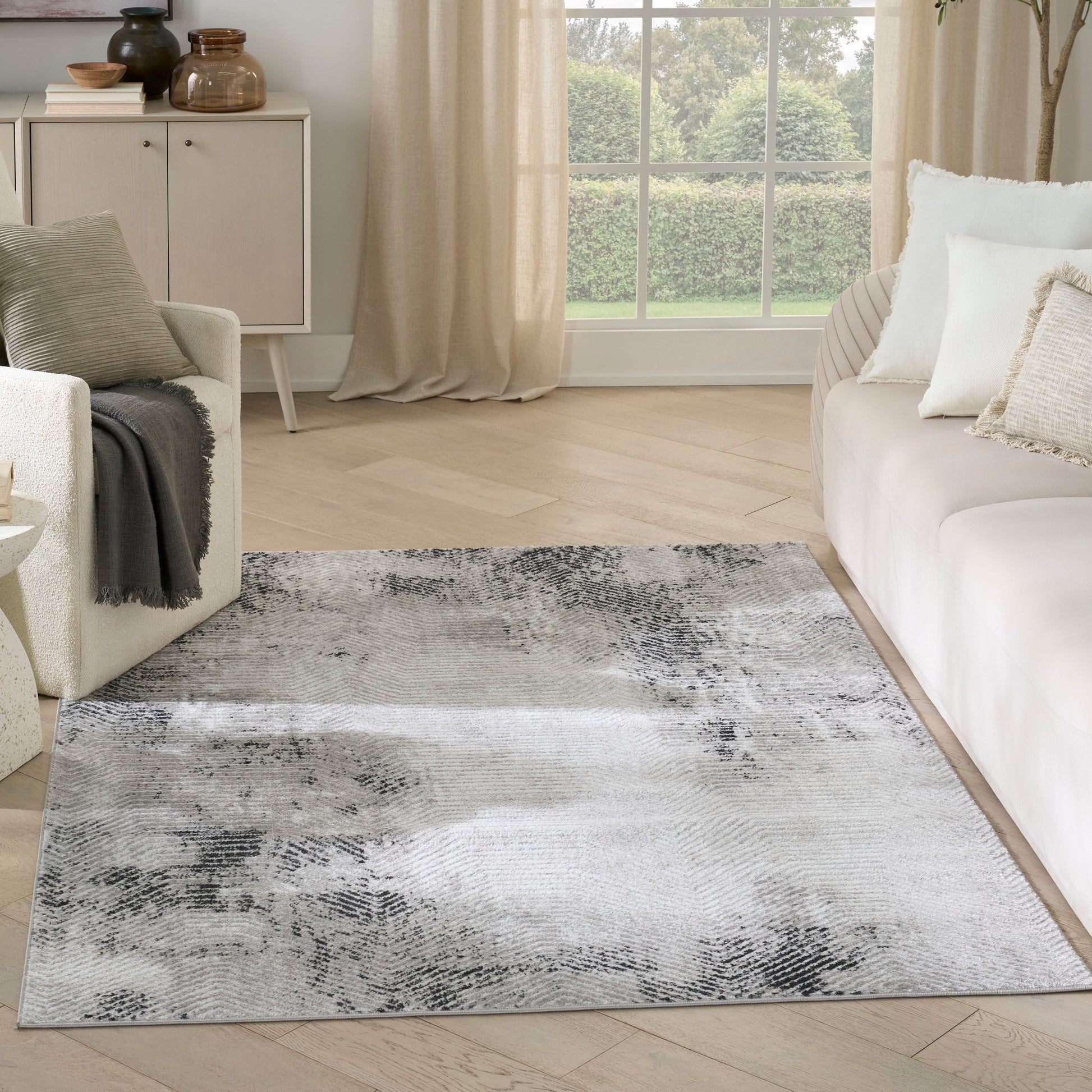 Abstract gray and ivory area rug over light wood floor in a neutral living room with beige curtains and white sofa.
