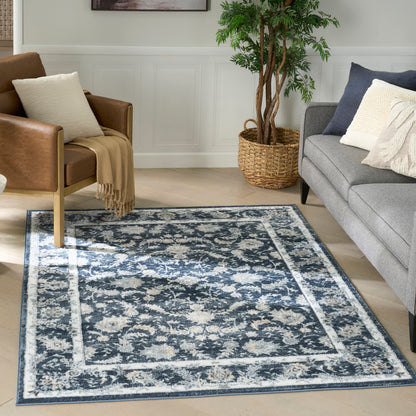 Blue distressed floral area rug with ivory border in living room; brown leather chair, gray sofa, potted plant.