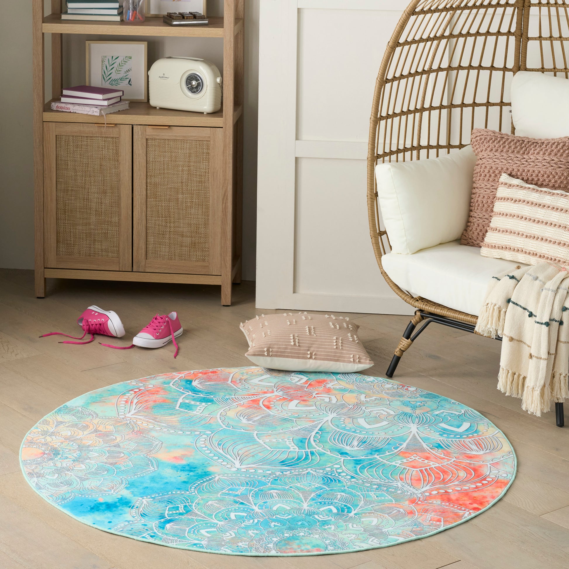 Round boho rug in turquoise, coral and teal with a white pattern, on light wood floor beside a wicker egg chair.