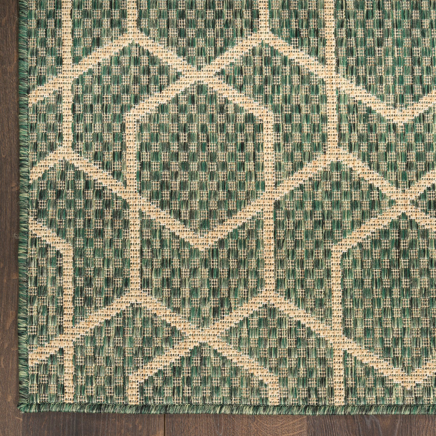 Green and cream woven rug with geometric diamond/trellis pattern; fringed edge, on dark wood floor.