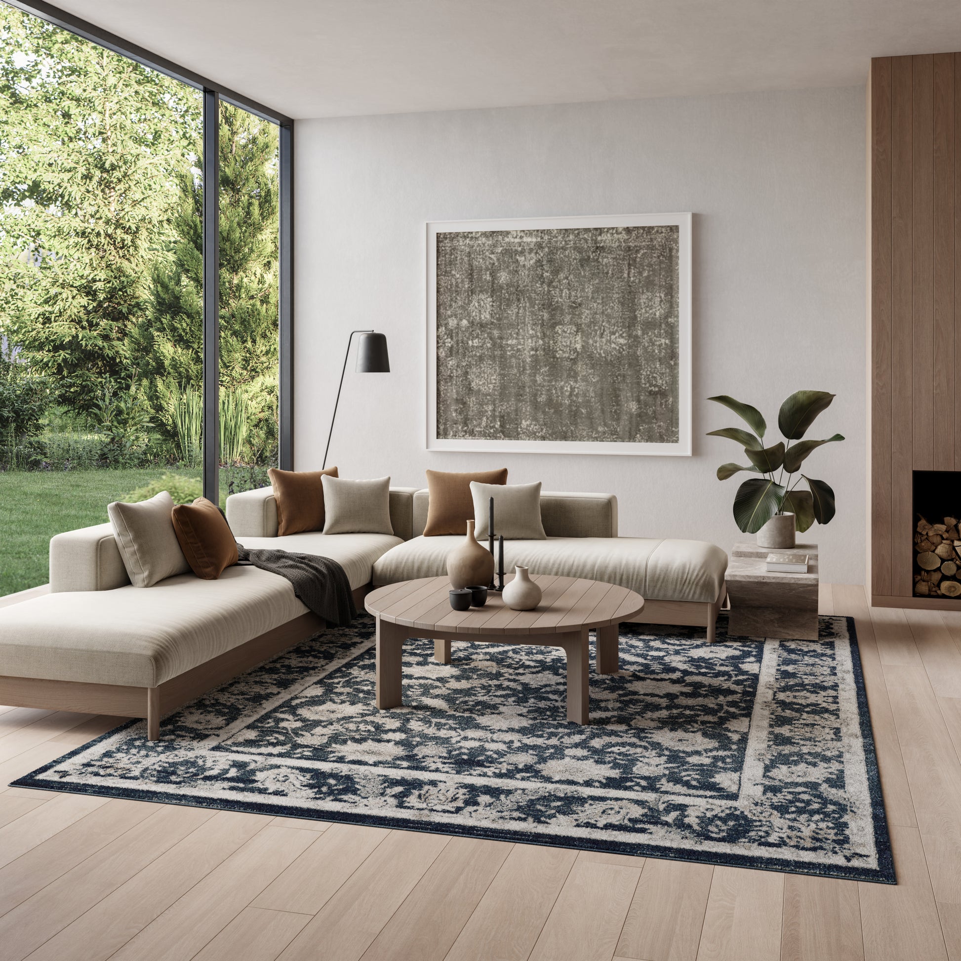 Neutral L-shaped beige sofa with brown and cream cushions, round wooden coffee table, blue patterned rug, large windows.