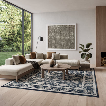 Neutral L-shaped beige sofa with brown and cream cushions, round wooden coffee table, blue patterned rug, large windows.