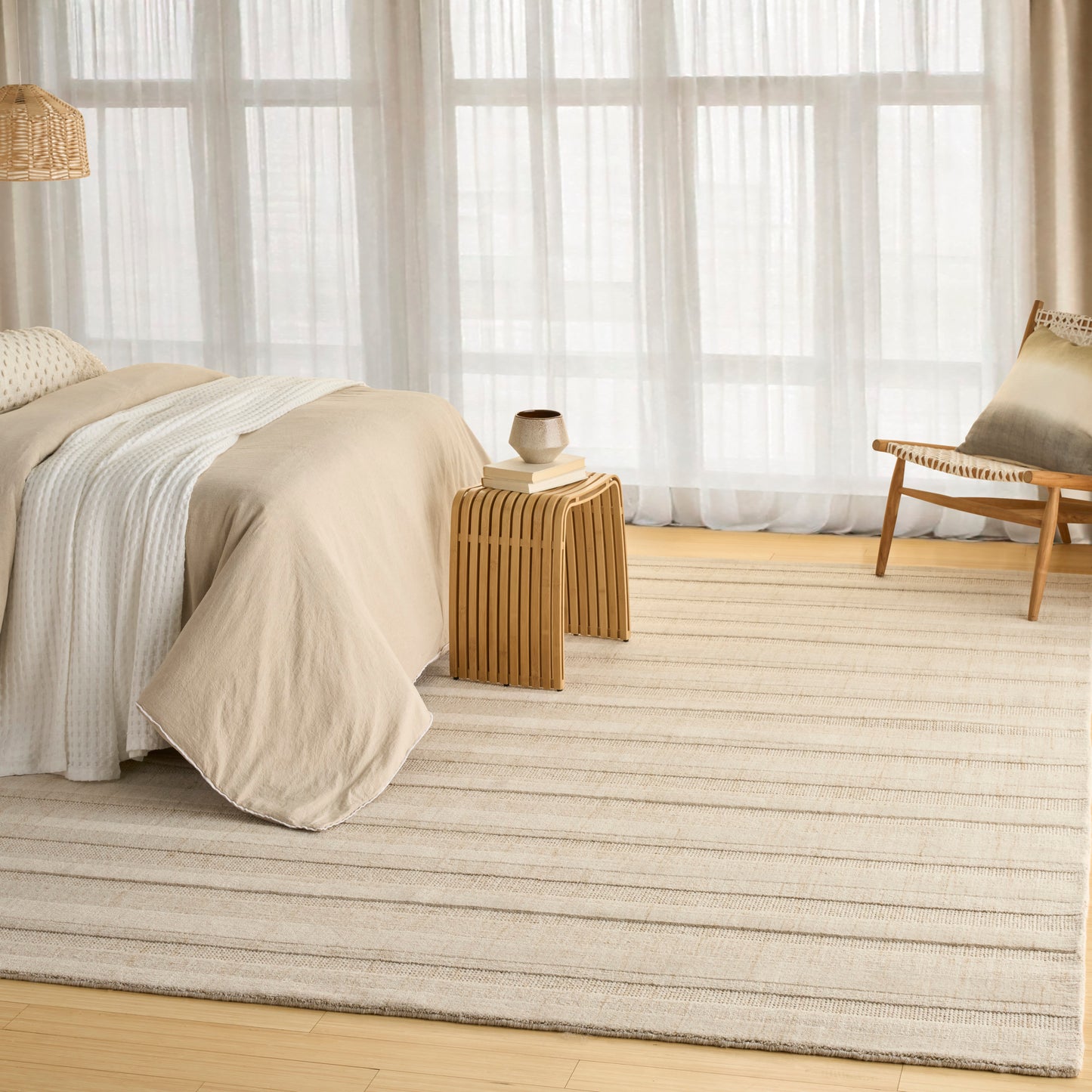 Neutral beige bedroom with linen-style bedding on a bed, striped cream rug, wooden side table and chair, sheer curtains.